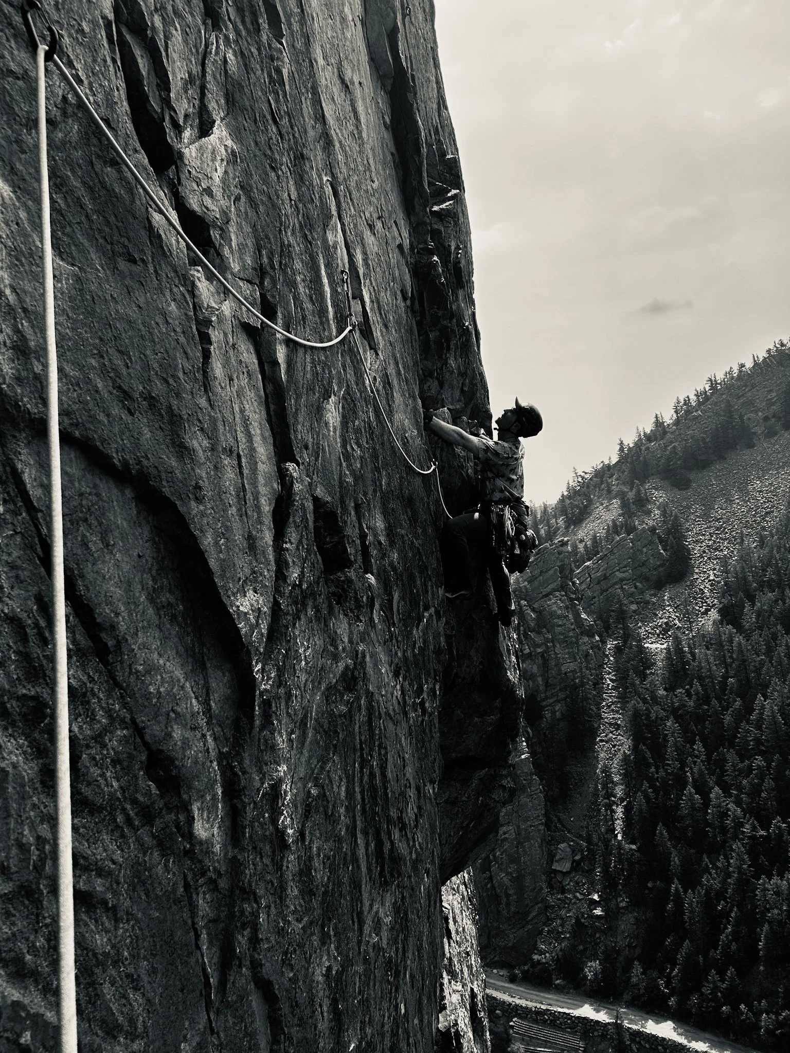 A person mountain climbing on a steep rocky cliff, wearing a helmet and harness, with a safety rope attached to the rock face, in a mountainous landscape with trees and a river below.