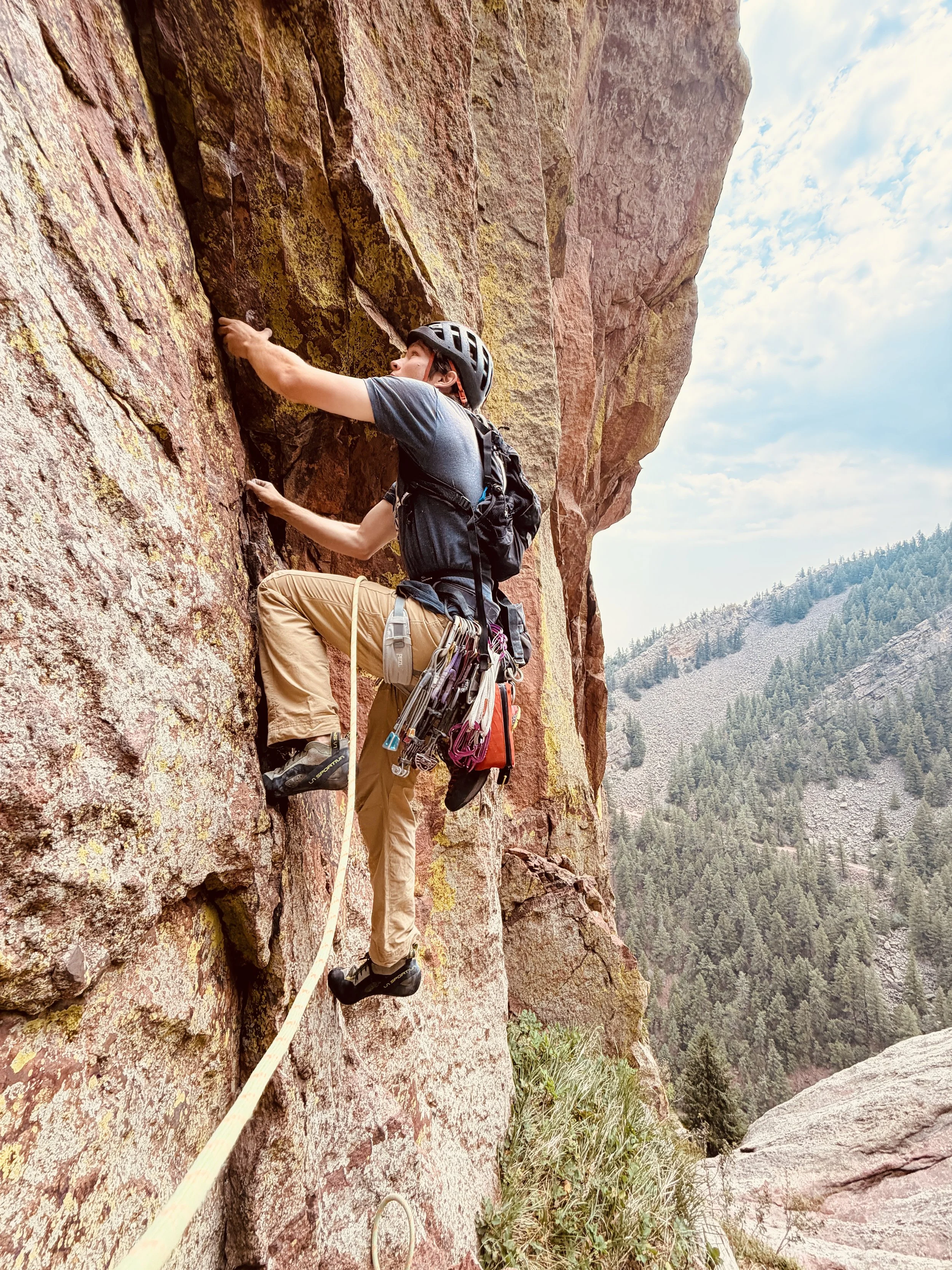 A person rock climbing on a steep, reddish-bink cliffside in a mountainous area. They are wearing a helmet, harness, and outdoor clothing, and are holding onto the rock with both hands. The background shows a mountain range and partly cloudy sky.