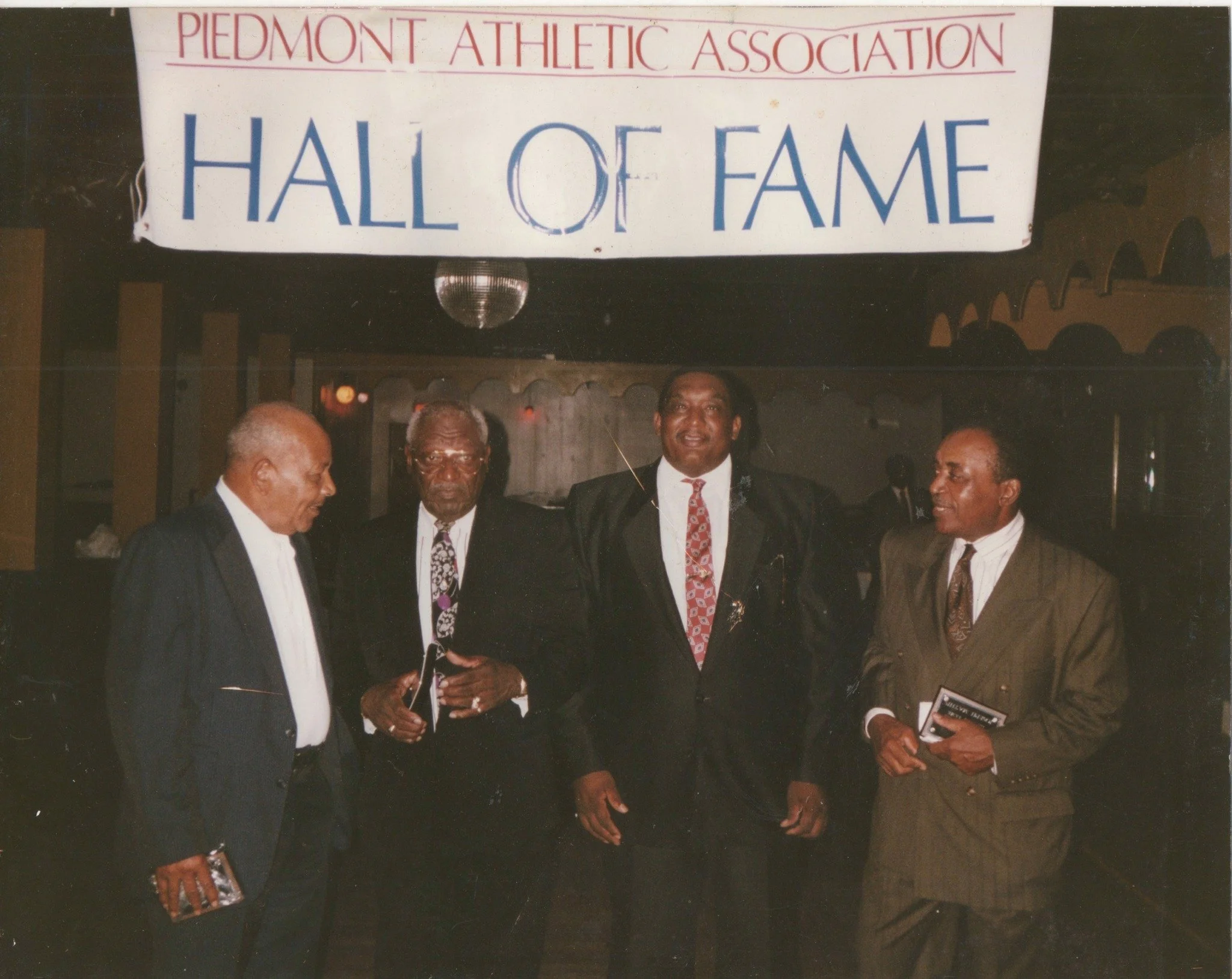 A group of five men dressed in formal suits standing together at an indoor event, with a sign above that reads 'Piedmont Athletic Association Hall of Fame.'