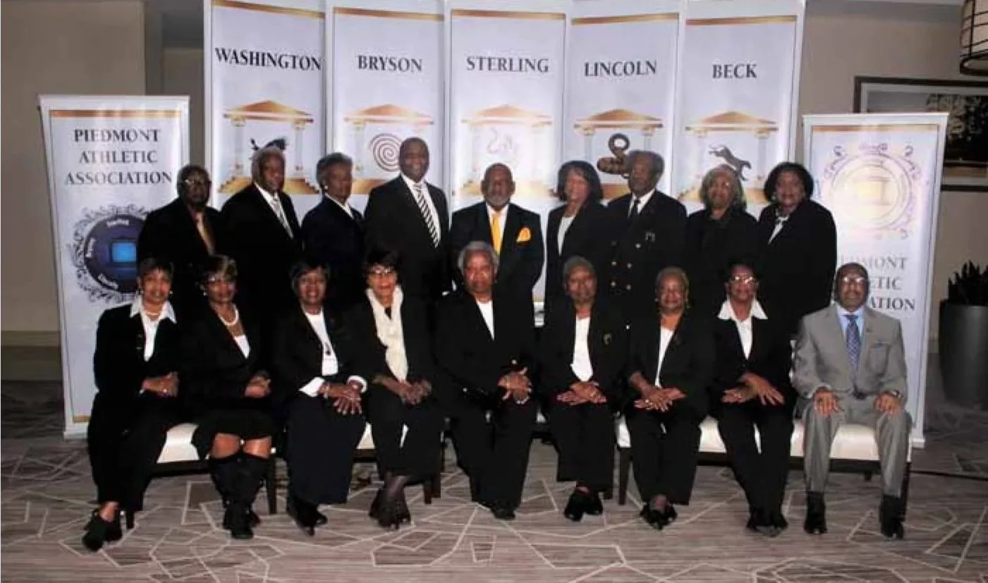 A group of 16 formally dressed individuals, mostly women in black suits and men in suits, posing for a photo in front of banners with various names and logos, including Washington, Bryson, Sterling, Lincoln, Beck, and Piedmont Athletic Association.