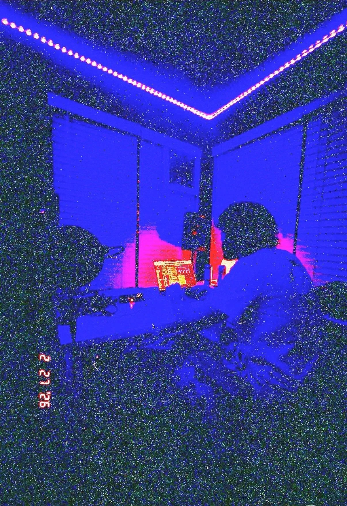 Room illuminated with blue and pink LED lights, two people with curly hair sitting at a desk with a monitor, speakers, and a laptop. Window with closed blinds in the background, reflecting the colorful lighting.