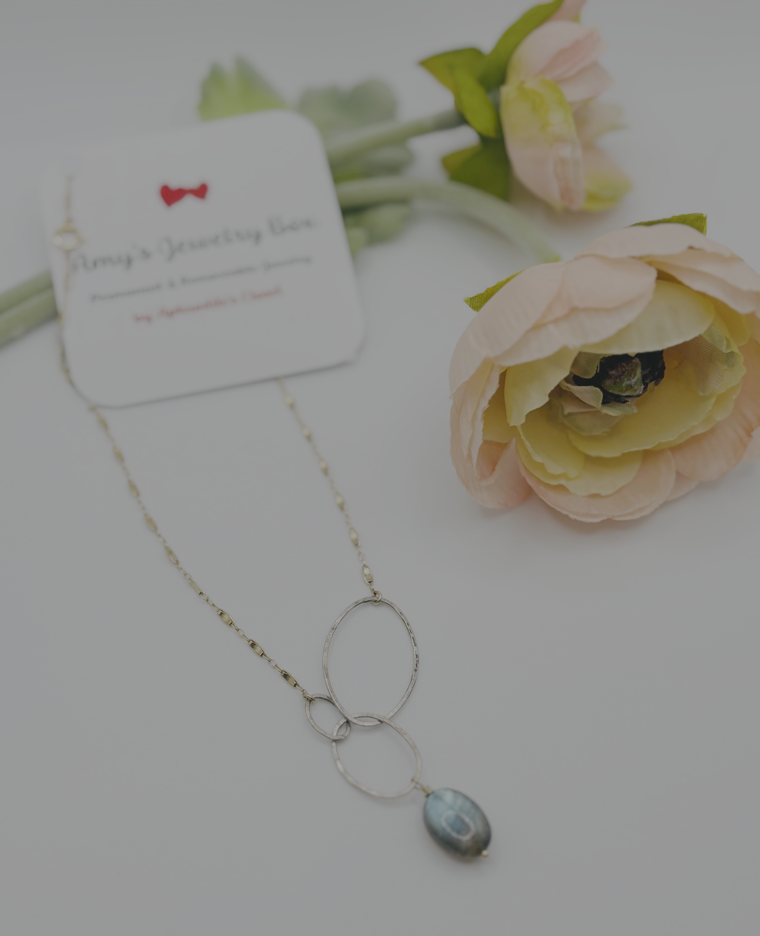 Close-up of a layered necklace with moonstone pendant, lying next to pink and cream artificial flowers and a white card with a red bow and partial text.