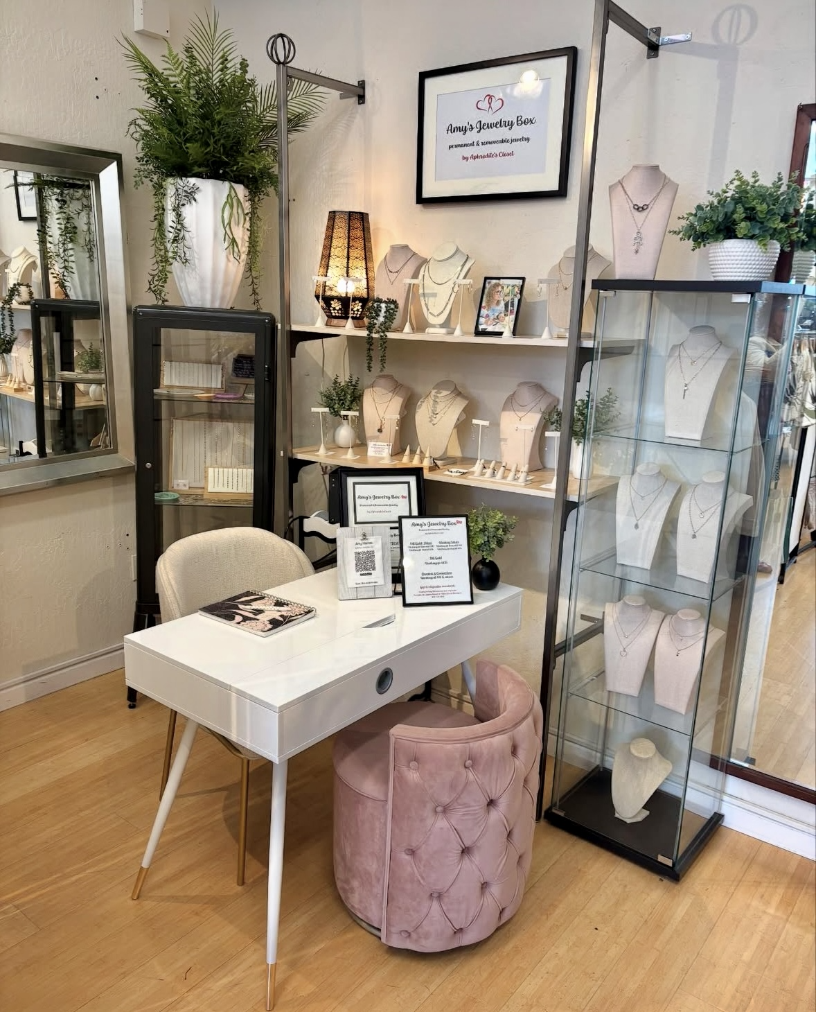 A jewelry boutique display with necklaces on mannequin busts, a glass cabinet with jewelry, a white table with promotional materials, a pink cushioned chair, and a black display case. The background has wall-mounted shelves with jewelry and a framed sign that reads 'Amy's Jewelry Box - Purseament & Reusable Jewelry by Ambitious Co.