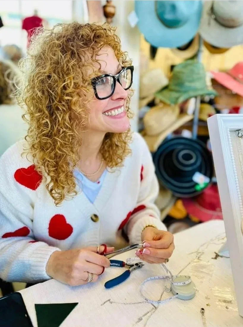 A woman with curly red hair, wearing glasses and a white sweater with red hearts, is smiling and working on jewelry at a craft table in a store that sells hats and accessories.