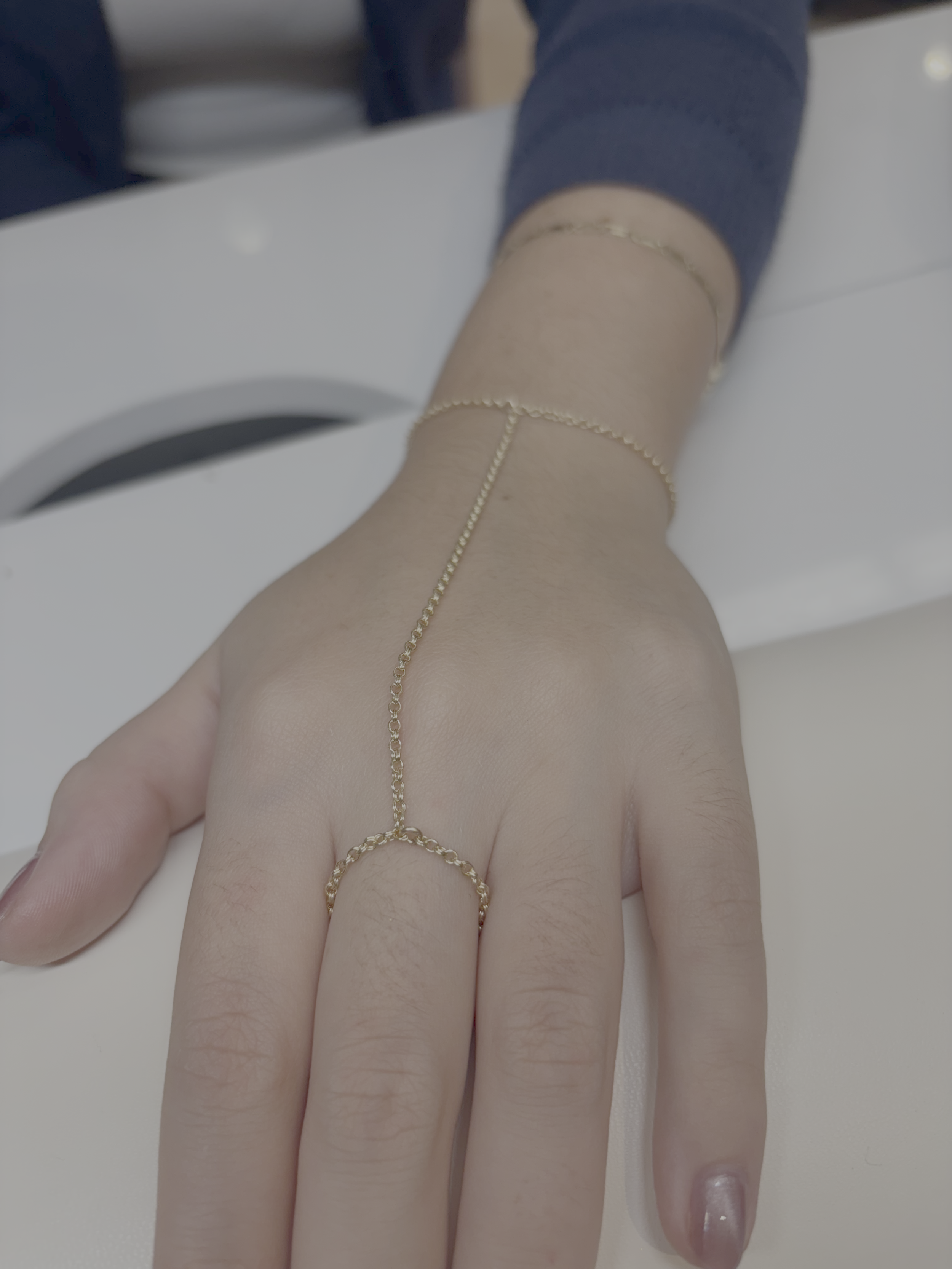Close-up of a hand with long fingers and well-manicured nails resting on a white surface. The hand is adorned with a delicate gold chain connected to a small ring on the middle finger. The person is wearing a dark long-sleeve shirt.