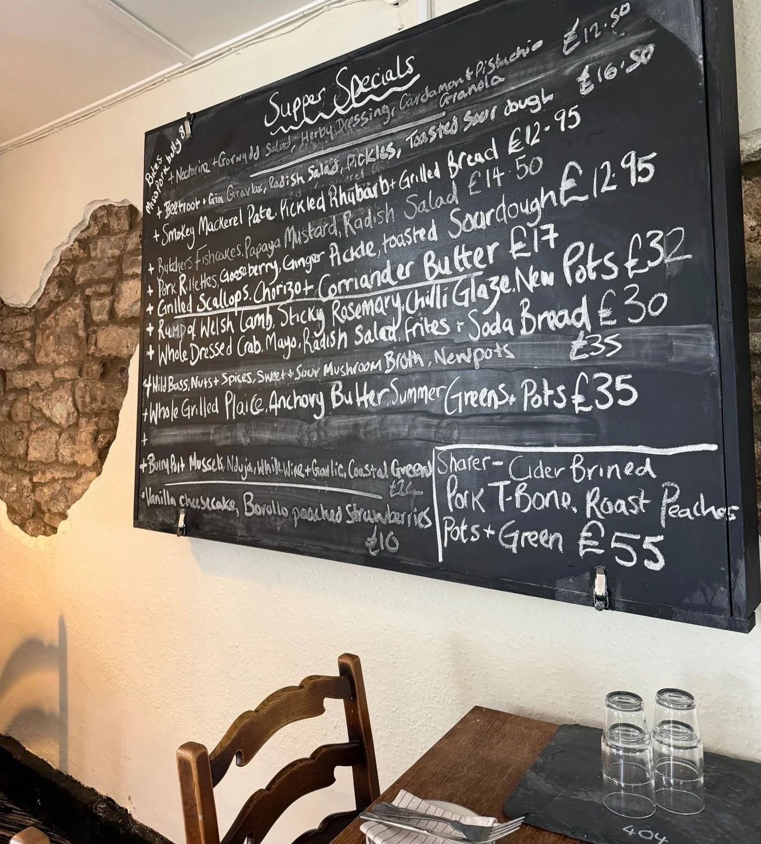 A traditional blackboard menu at The Butchers Arms displaying seasonal food and drink specials in white chalk, beside a neatly set wooden dining table in the pub's welcoming interior.