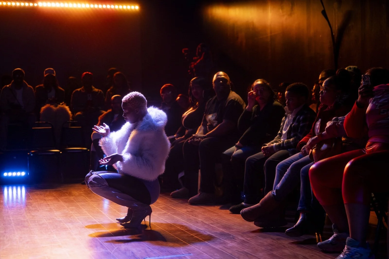 A performer dressed in a white fur coat and high heels is crouching on stage with one knee bent, while an audience watches from the darkened background. The performer is illuminated by colorful stage lighting, and the audience sits in a curved arrangement, watching attentively.