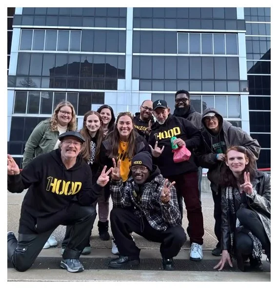 Group of ten people smiling and posing outside in front of a modern glass building, some making peace signs, wearing casual clothing and jackets.