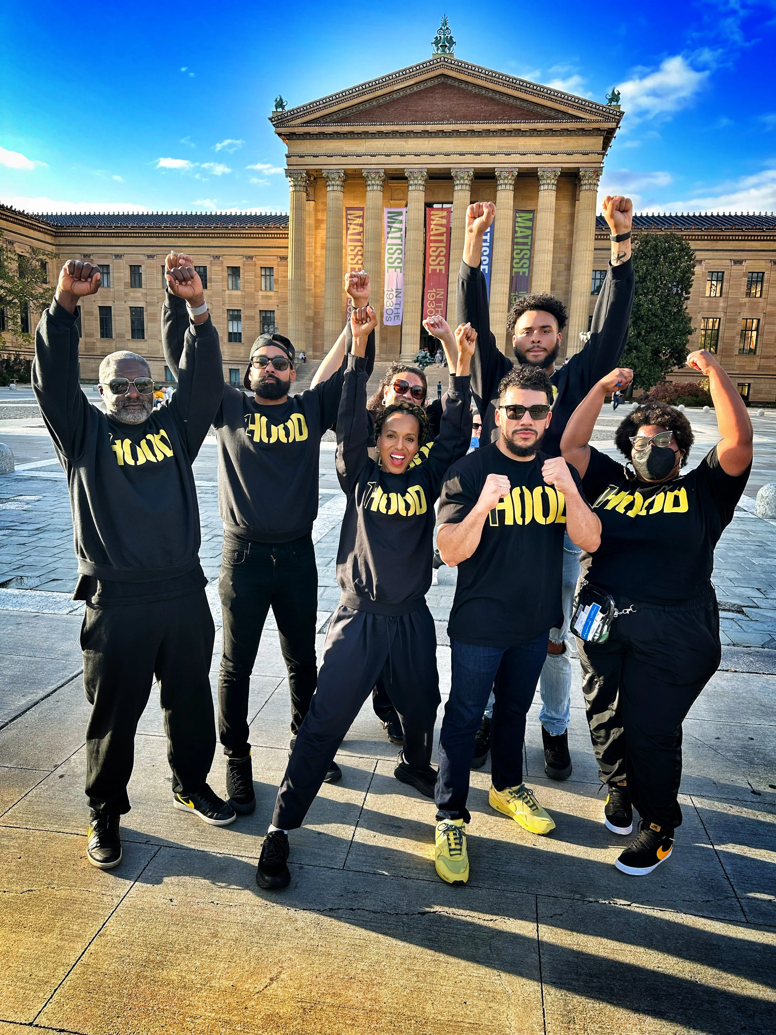 A diverse group of seven people, dressed in black hoodies with yellow text, poses with their fists raised in front of the steps of a historic building with columns, outside on a sunny day.