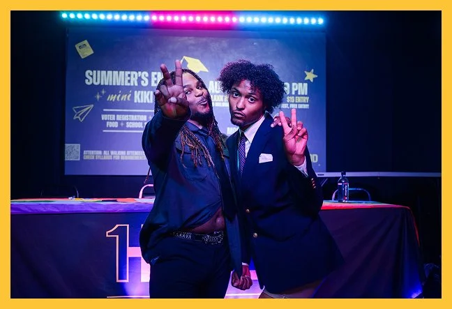 Two men standing on stage with a banner in the background. One man has dreadlocks and is wearing a dark shirt, while the other has curly hair and is dressed in a suit. Both are making peace signs and looking at the camera.