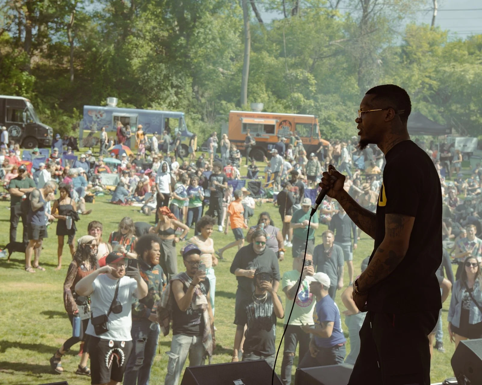 A performer on stage holding a microphone at an outdoor concert with a large crowd gathered on the grass, with food trucks and trees in the background.