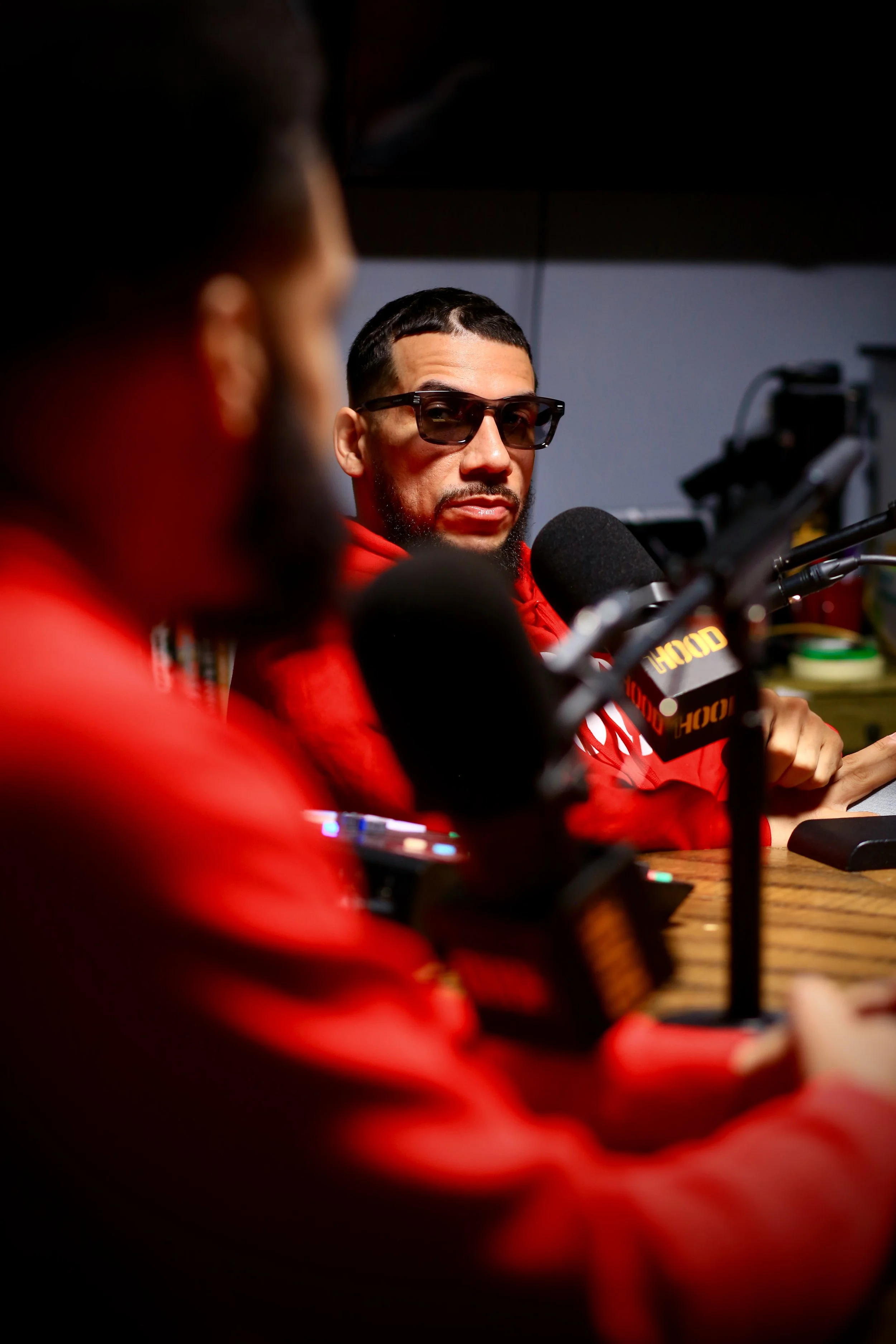 A man with short dark hair, beard, and sunglasses speaking into a microphone during a press conference or interview, wearing a red jacket. Another person, blurred in the foreground, is also wearing a red jacket. The background features a computer and electronic equipment.