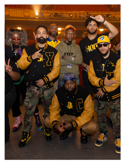 Group of six people in streetwear and varsity jackets posing together indoors. Some wear hats and sunglasses. The setting appears to be a stage or event space with warm lighting.