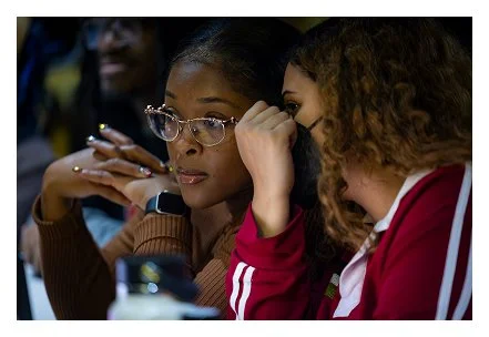 Two women sitting closely together, one wearing glasses and the other with curly hair, engaging in a conversation or listening.