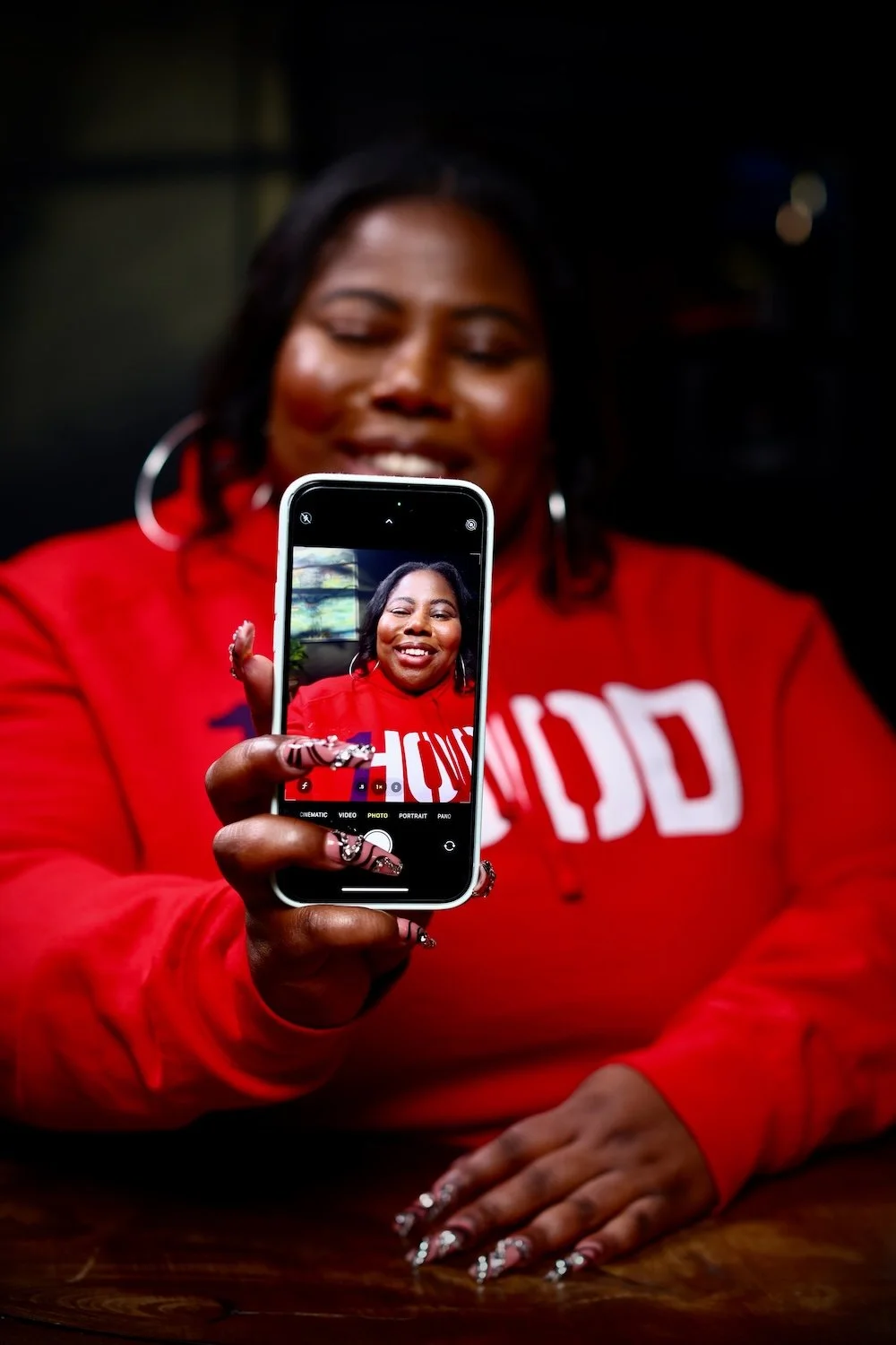 A woman in a red hoodie taking a selfie with a smartphone, smiling at the camera, with long painted nails and hoop earrings.