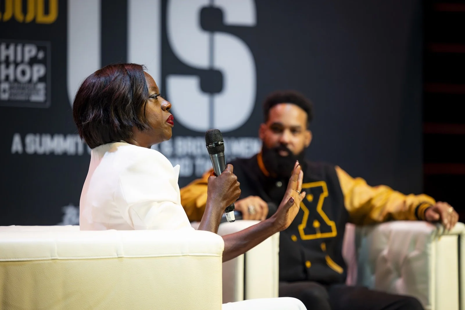 A woman in a white blazer speaking into a microphone during a panel discussion, with a man in a black and yellow jacket sitting beside her, on stage at a conference with a large logo and text in the background.
