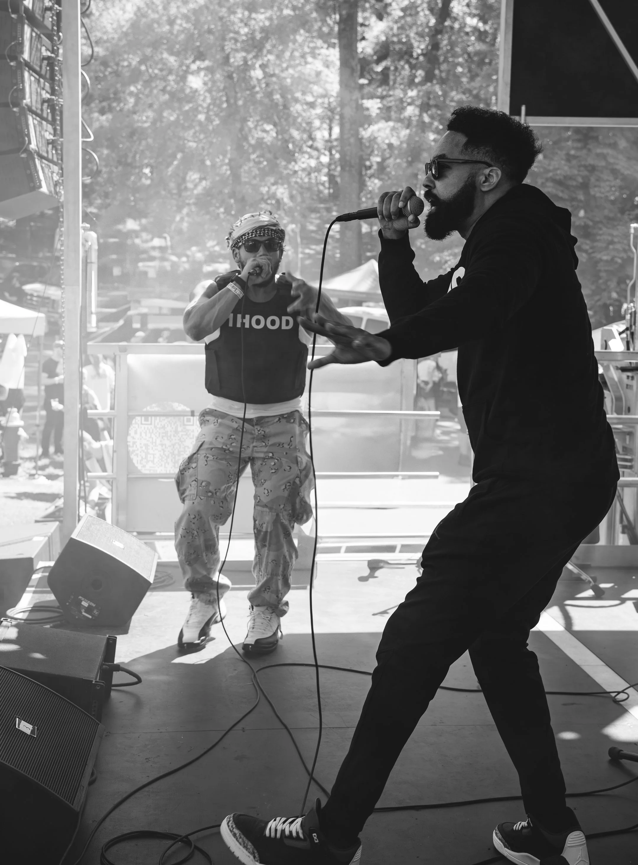 A man with a beard, glasses, and a hoodie is holding a microphone and singing or talking on stage. Another person with sunglasses, a patterned headscarf, and patterned pants is also holding a microphone and performing. The stage has various equipment, and trees are visible in the background.