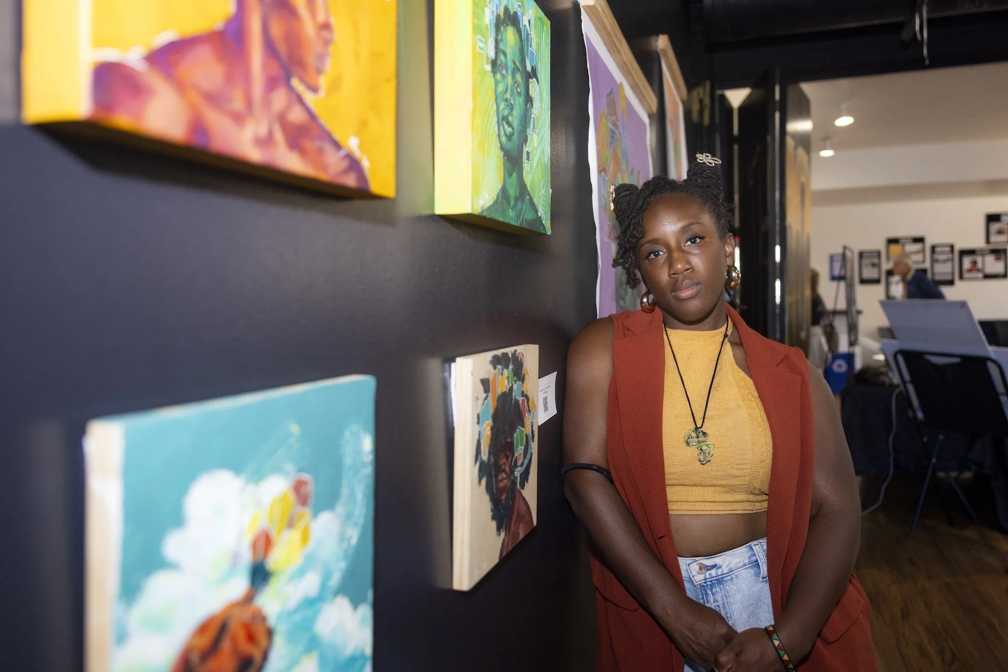 A woman with dark skin and braided hair tied into two buns, wearing large gold earrings and a necklace with an African continent pendant, stands in art gallery near colorful paintings on a black wall.