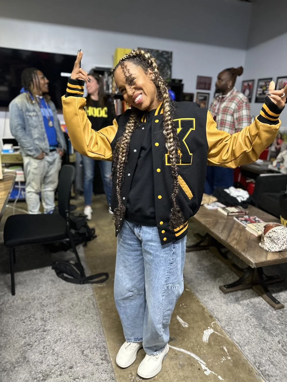 A woman with braided hair smiling and making peace signs with both hands, wearing a black and yellow varsity jacket, light blue jeans, and white sneakers, in a room with other people and tables in the background.