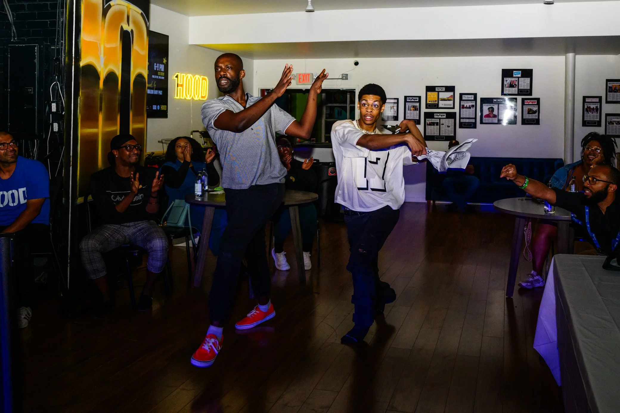 A group of people inside a restaurant or lounge, with two individuals standing and dancing in the middle, others sitting and clapping around them. The setting has framed pictures on the wall and a neon sign that reads 'HOOD'.