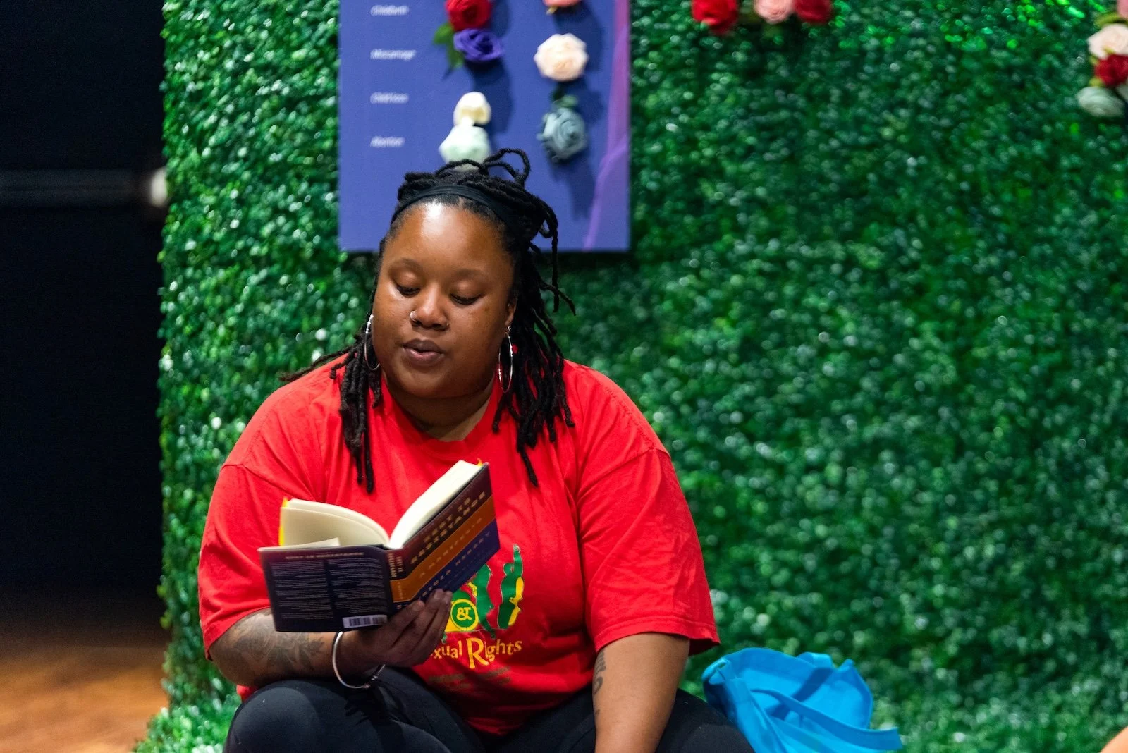 A woman wearing a red t-shirt and earrings, sitting in front of a green wall, reading a book.