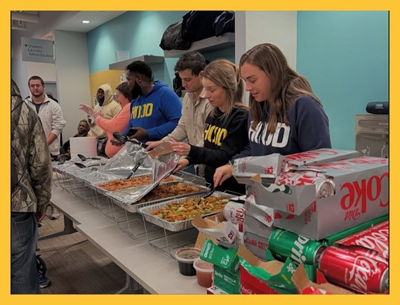 People in line at a food service counter with trays of food, pizza boxes, and soft drinks.