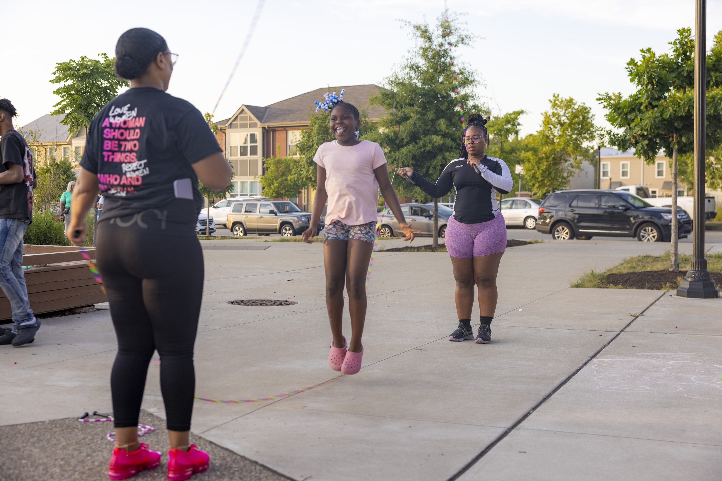 A girl with a blue and white bow hair accessory jumping rope while smiling. Other women are in the background, one holding the jump rope. It is an outdoor setting with trees, parked cars, and residential buildings.