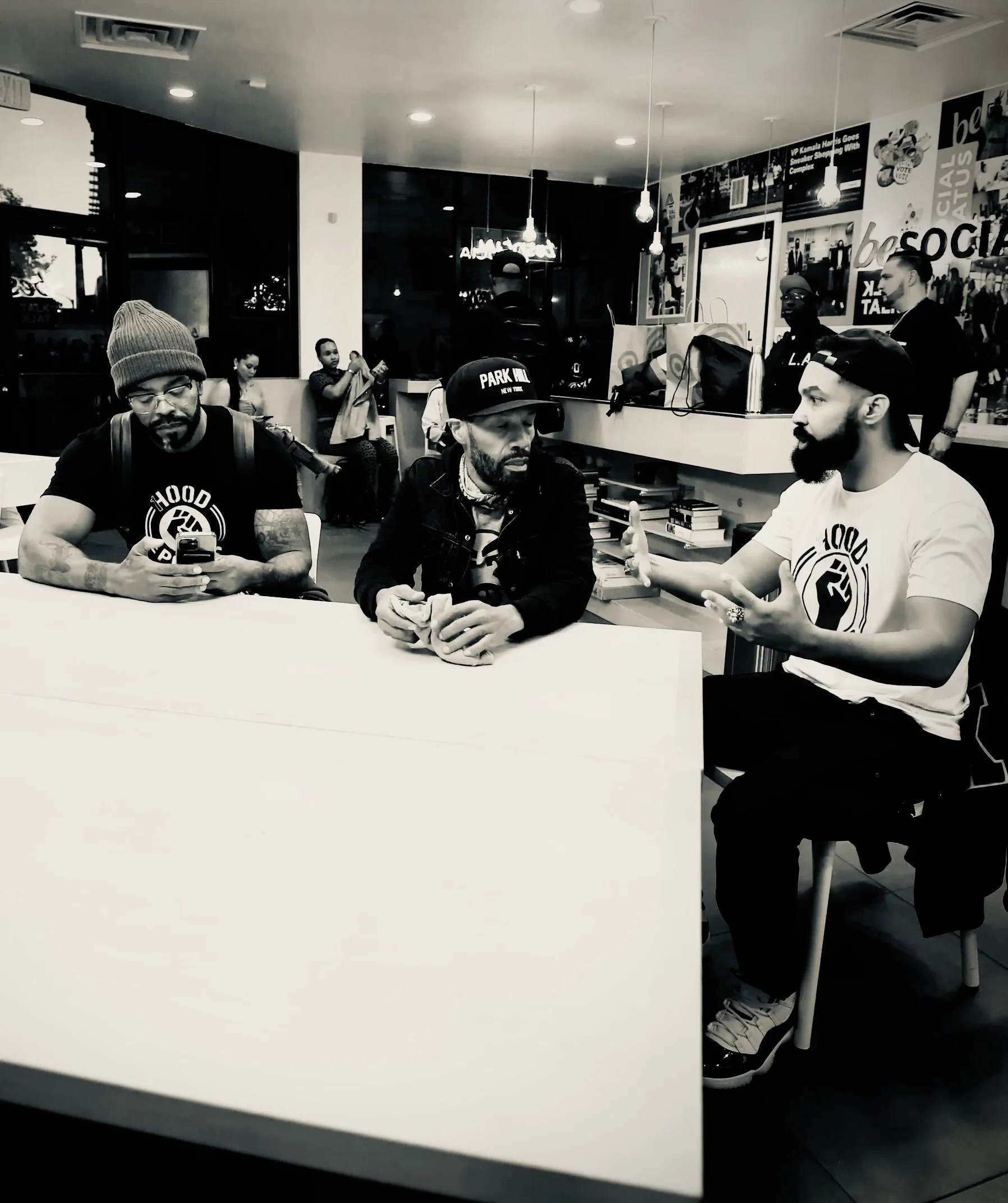 People sitting at a table in a modern fast food restaurant with posters on the wall, some using phones and engaged in conversation, black and white photo.