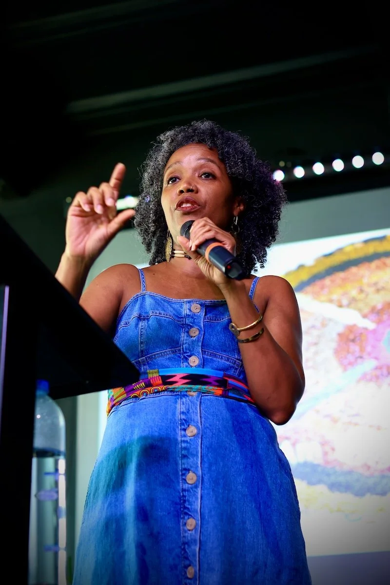 A woman with curly black hair, wearing a blue denim dress, holding a microphone, gesturing with her left hand, standing in front of a projected colorful landscape image.