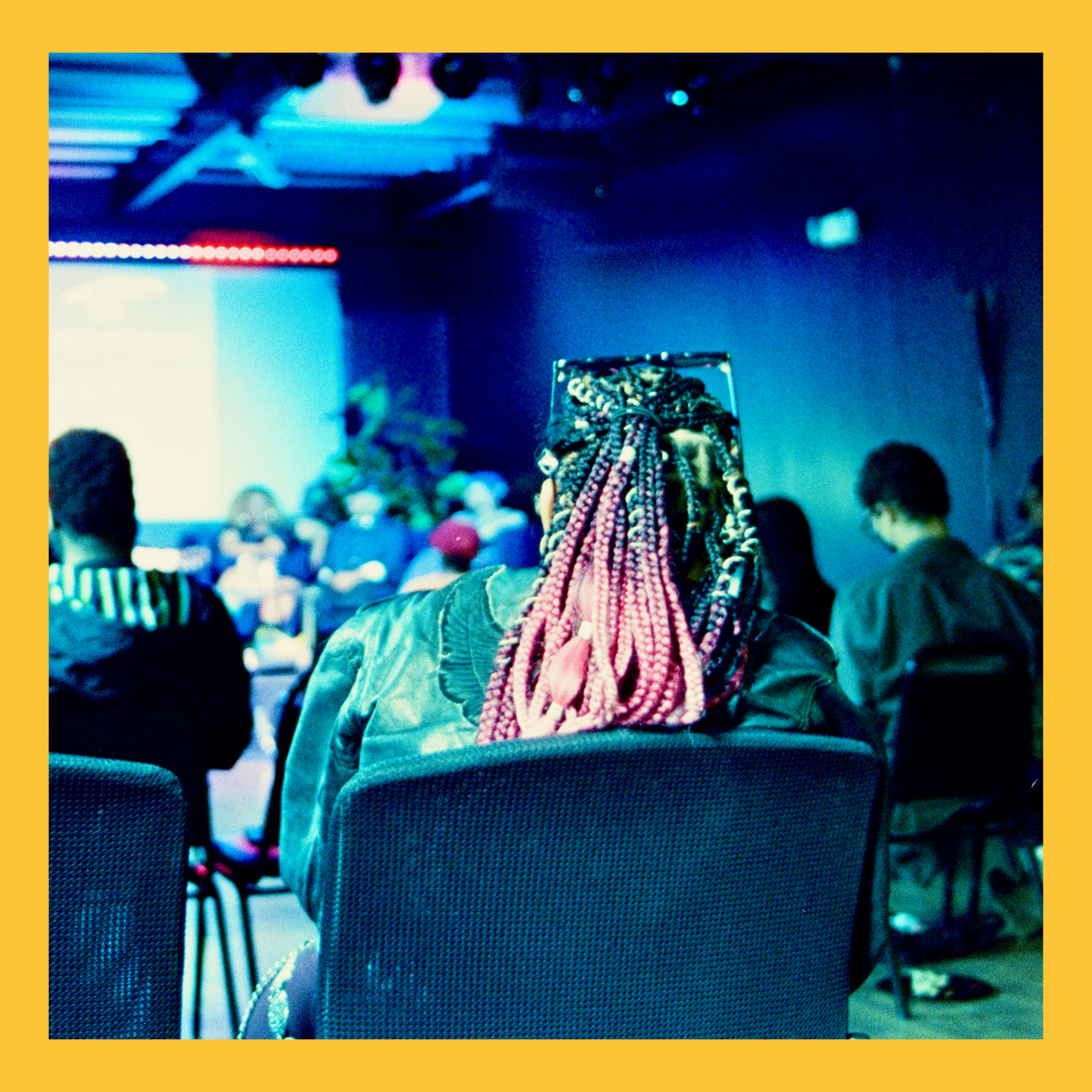Person with braided hair sitting in a blue-lit room, possibly a nightclub or bar, with people and bicycles blurred in the background.