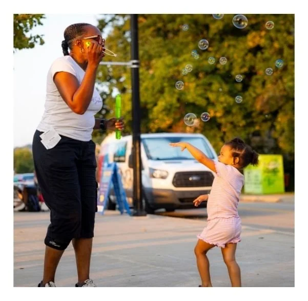 A woman making bubbles with a bubble wand and a young girl reaching out to catch bubbles in an outdoor park or street setting.