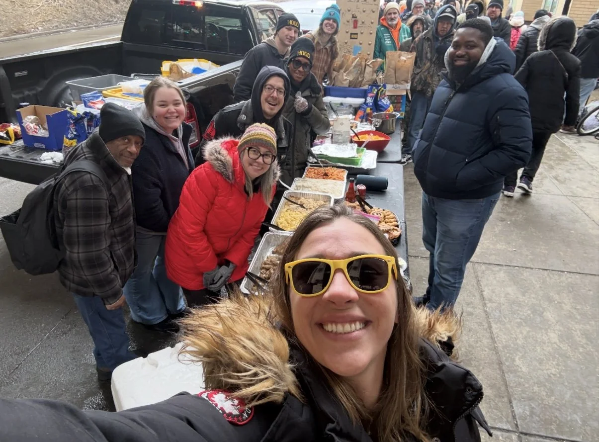 Group of people smiling and standing behind a table with food at an outdoor gathering, dressed in winter clothing.