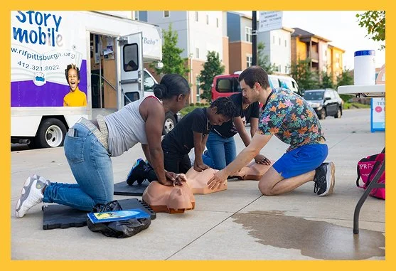 Group of four people practicing CPR on mannequins outdoors near a mobile health van.