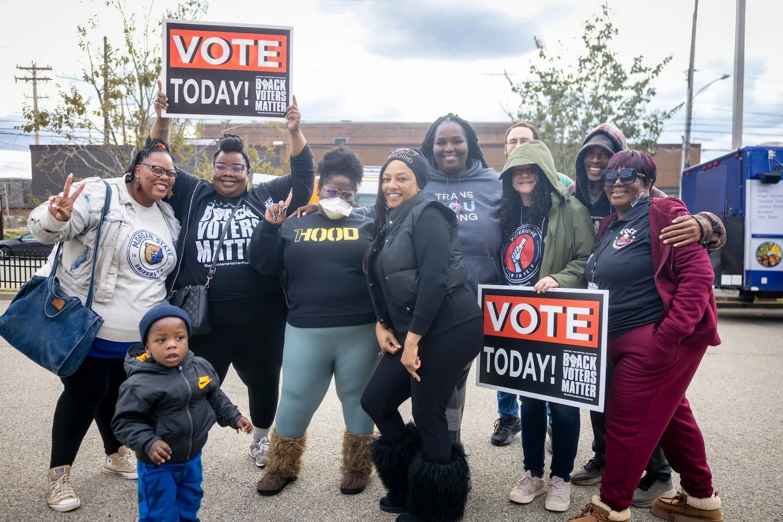 Group of diverse people standing outdoors, holding signs that say 'Vote Today! Back Voters Matter,' smiling, with a small child in the foreground and a food truck in the background.