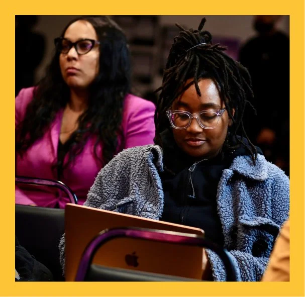 Two women sitting in an audience, focused on something in front of them. The woman in the foreground has glasses and is wearing a blue jacket. The woman in the background has glasses and is wearing a pink blazer.