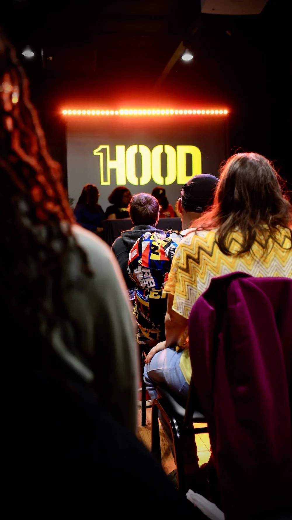 Audience watching a presentation on stage with a screen displaying the word 'HOOD' in yellow letters, illuminated by red lights.