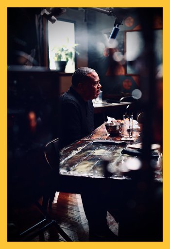 An older man sitting alone at a dark table in a dimly lit room with natural light coming through a window, with a bowl and a glass on the table, and a microscope or camera mounted overhead.