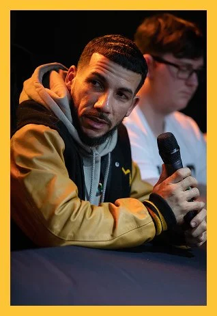 A man with a beard and short hair, wearing a jacket and hoodie, holding a microphone and speaking at a panel or discussion event, with another person blurred in the background.