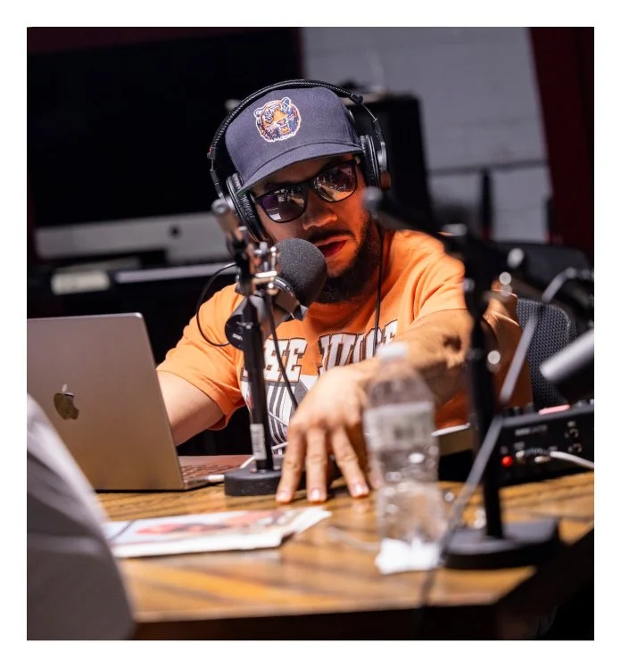 Man wearing sunglasses, a baseball cap with a logo, and headphones, speaking into a microphone in a recording studio, with a laptop and water bottle on the table.