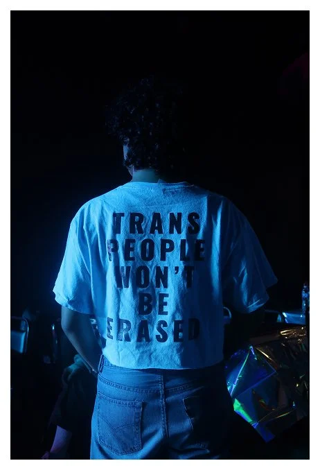 Person wearing a white T-shirt with the message 'Trans People Won't Be Erased' printed on the back, standing in a dark environment.
