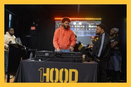A DJ with curly hair and a beard wearing an orange hoodie performs at a DJ booth with 'HOOD' written on the front, while several people observe in a dark room with colorful lights.