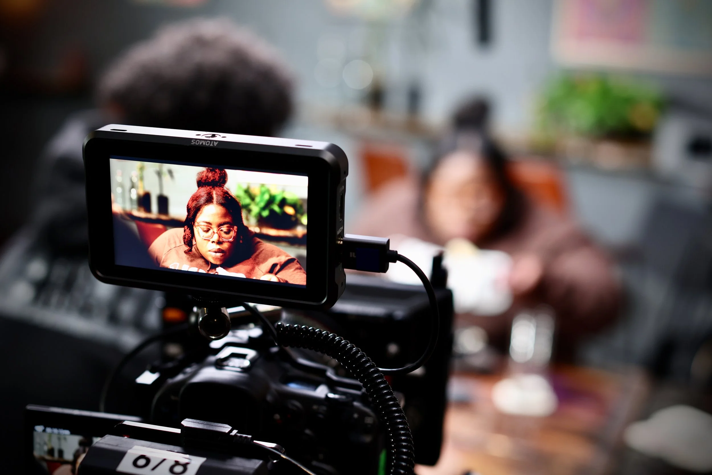 Close-up photo of a professional camera monitor capturing a woman who is in focus on the monitor. The background is blurred, indicating an indoor setting.