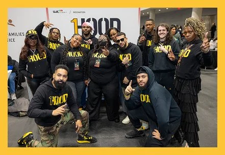Group of diverse people posing together at an event, some making fists and pointing, wearing black hoodies with yellow text, in front of a backdrop with a 100D logo.