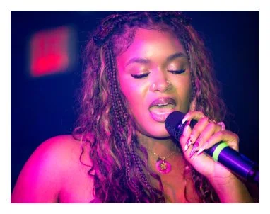 A young woman singing into a microphone on stage with pink and purple lighting.
