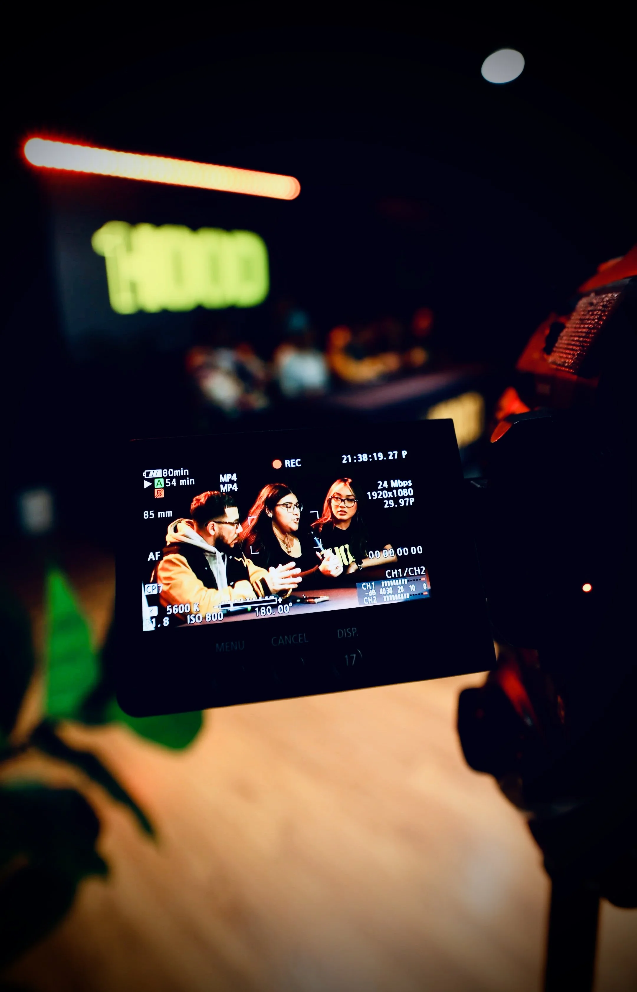 A video camera's display showing three people sitting at a table engaged in conversation, with a recording timestamp, technical camera settings, and a blurred background of a scene with a lit sign.