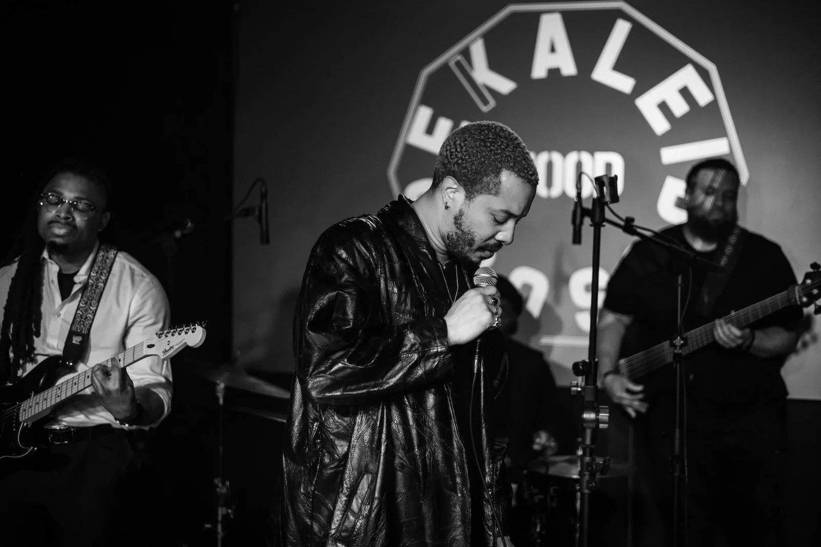 Musicians performing on stage, with a man singing into a microphone in the foreground and two guitar players on either side, in front of a backdrop with the text 'KALEIDOSCOPE'.