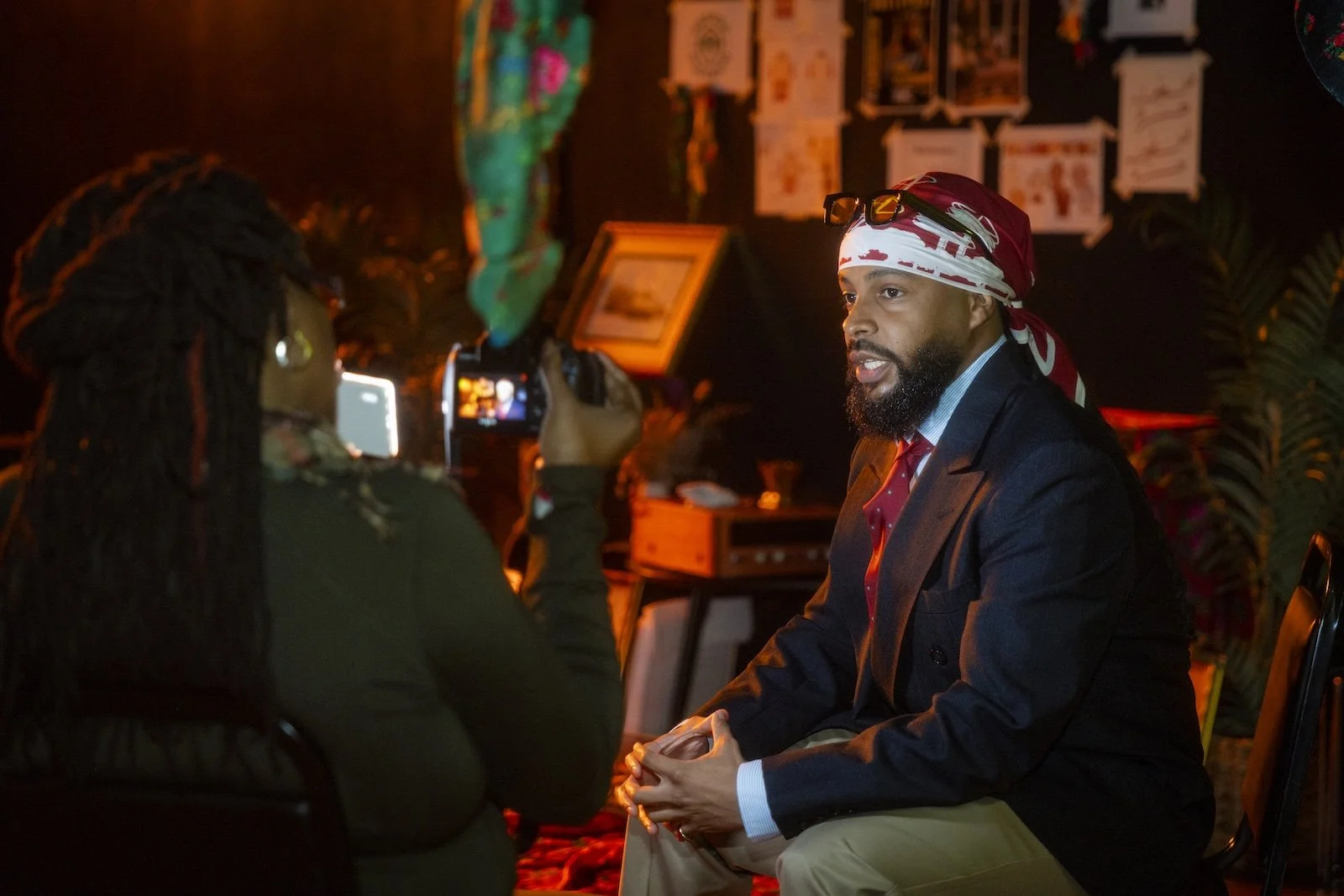 A man in a suit who is being interviewed by a woman holding a camera. They are sitting indoors in a dimly lit room with various artworks and a potted plant in the background.