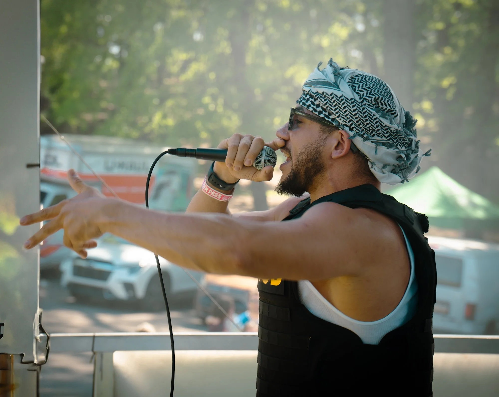 A man with a beard, sunglasses, and a patterned headscarf speaking into a microphone during an outdoor event, with trees and tents in the background.