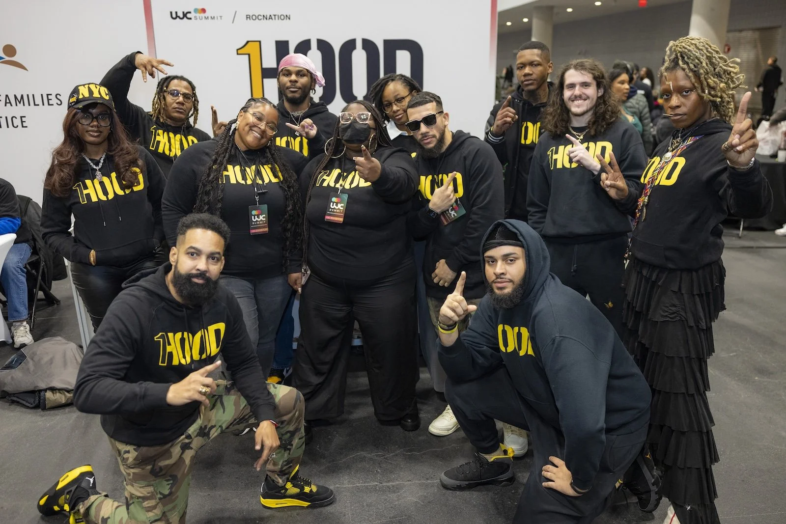 Group of diverse individuals posing at a conference, all wearing black hoodies with the word 'HOOD' in bold yellow letters, some making hand gestures. Background shows a large banner with logos and event information, indicating a professional or community gathering.