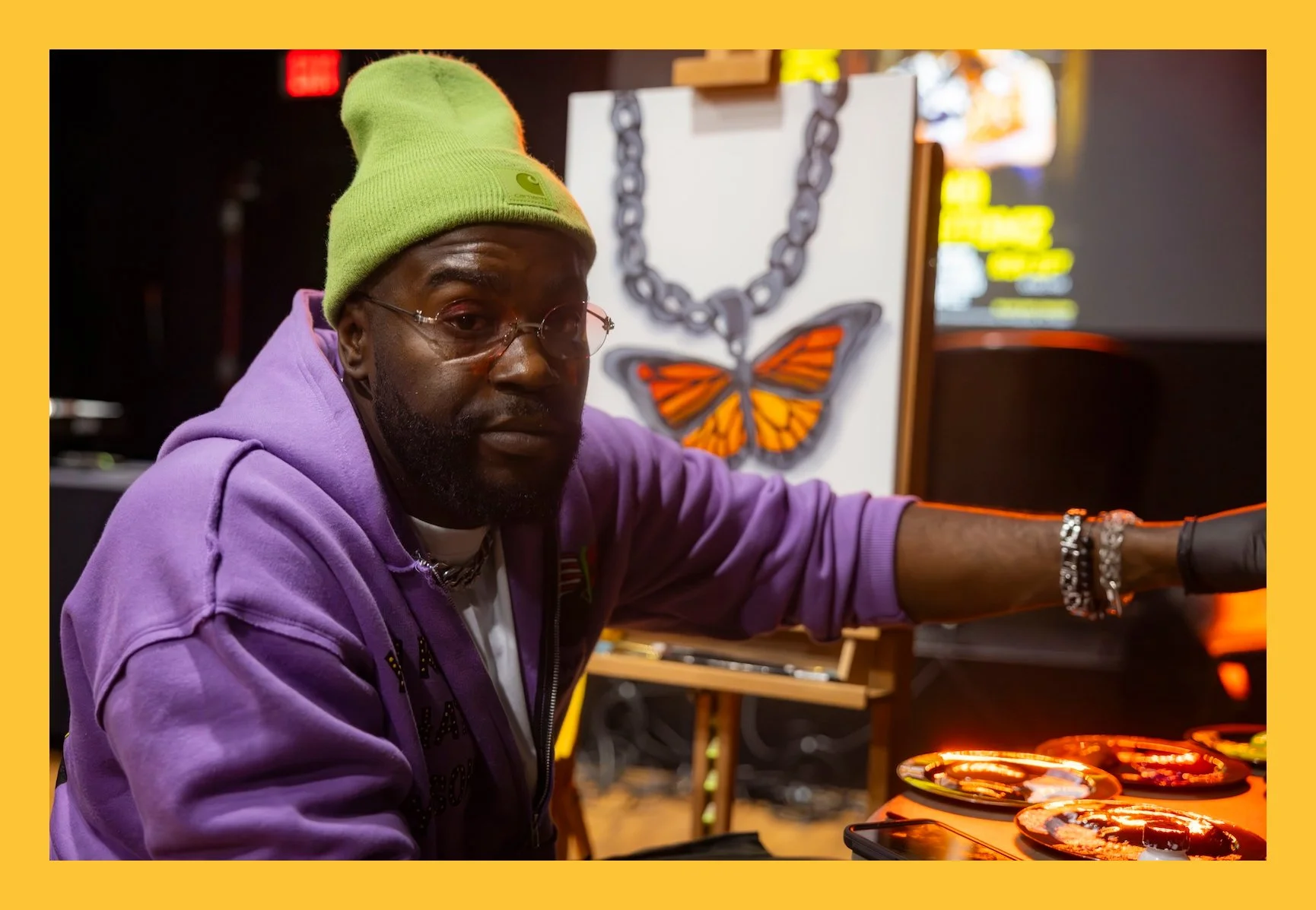 A man wearing a green beanie and purple hoodie, sitting in front of a computer screen with glowing artwork of a chain and butterfly. He has glasses and jewelry, and his arm is extended towards a control surface with illuminated buttons or pads.